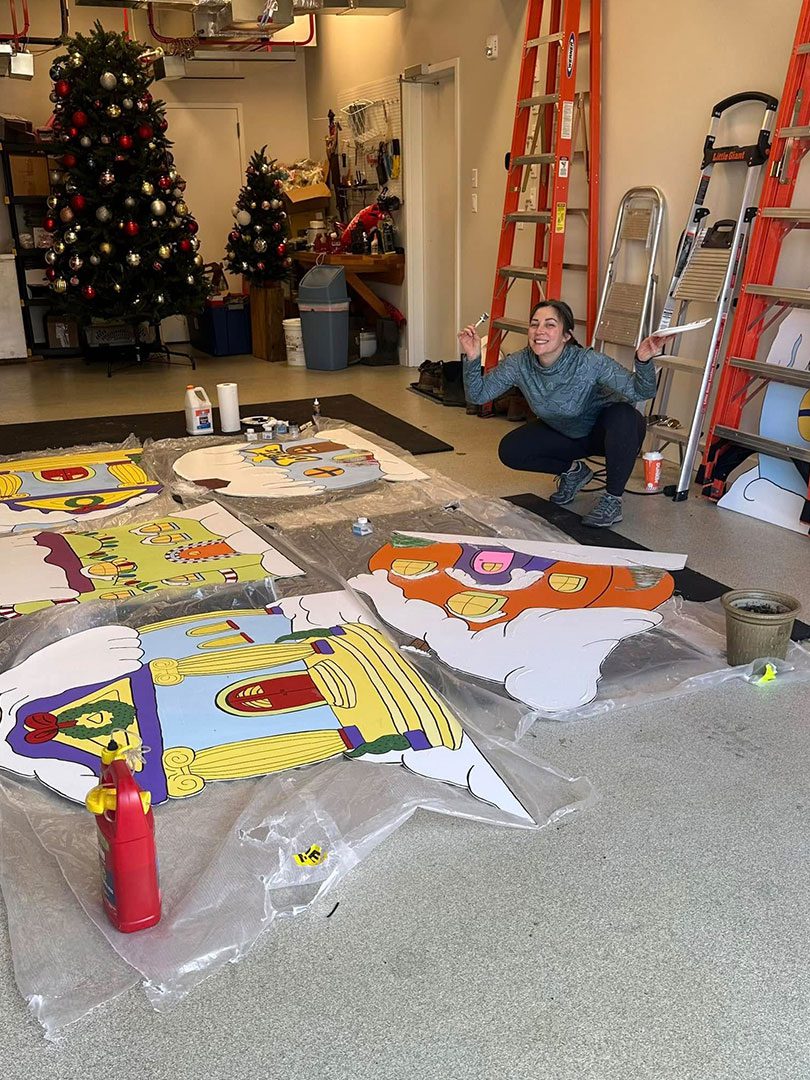 female staff member smiling and posing while painting large pieces for Christmas parade float female staff member smiling and posing while painting large pieces for Christmas parade float